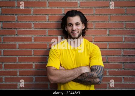 Fröhlicher männlicher Athlet mit kräftigem Körper und im Sportswear Stehen Mit gekreuzten Armen in der Nähe einer Ziegelwand und Blick auf die Kamera Stockfoto