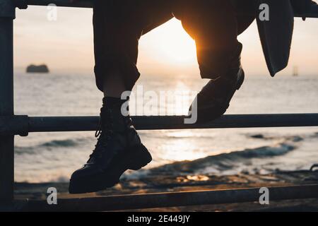 Crop anonyme Reisende in warme Kleidung und Stiefel sitzen auf Zaun Sie gegen winkende Meer und Sonnenuntergang über Horizont während Erholung am Meer im Herbst Stockfoto