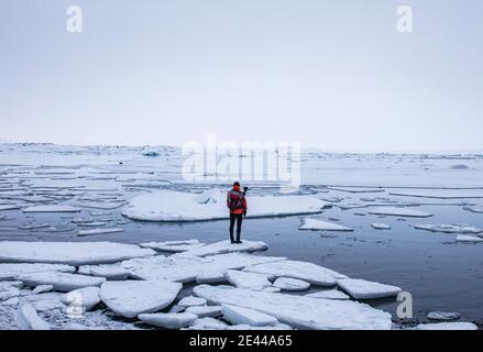 Rückansicht des anonymen Entdeckers in warmer Kleidung, die auf steht Stück Eis im Meer in der Nähe von Ufer und bewundern Winter seascape in Island Stockfoto