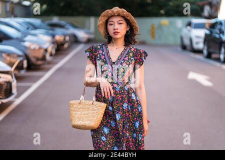 Asiatische Frau in Strohhut und mit Weidenhandtasche stehend Auf dem Parkplatz und Blick auf die Kamera Stockfoto