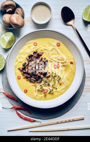 Draufsicht auf Schüssel mit appetitanregenden Sahnesuppe mit Nudeln Und gehackte Pilze auf dem Tisch mit Essstäbchen und frisch angeordnet Zutaten in asiatischen Restau Stockfoto