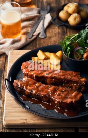 Hoher Winkel von appetitlichen Schweinerippen mit Grillsauce auf Teller mit rustikalen Kartoffeln und Salat serviert auf dem Tisch in Café Stockfoto