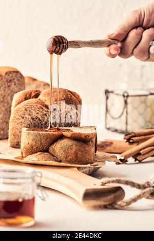 Ernte unkenntlich Person mit Dipper Gießen süßen Honig auf frisch Brot auf Schneidebrett auf dem Tisch in der Küche Stockfoto