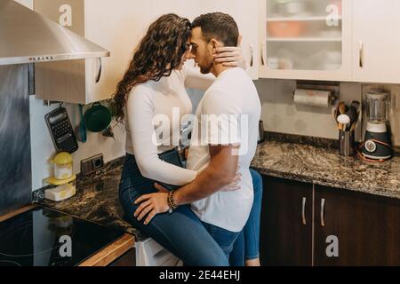 Lächelnde Frau, die auf der Theke in der Küche sitzt und ihren Freund umarmt, während sie sich gegenseitig anschaut Stockfoto