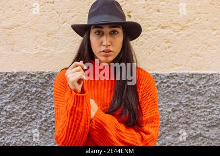 Ernst junge ethnische weiblich in leuchtend orange gestrickt Pullover und Stilvoller Hut, der die Kamera anschaut, während er gegen eine Ziegelwand steht Stockfoto