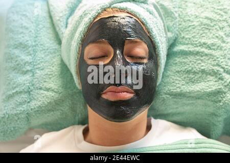 Nicht erkennbare junge weibliche Klientin in legerer Kleidung mit Handtuch auf Kopf und schwarze Tonmaske im Gesicht Stockfoto