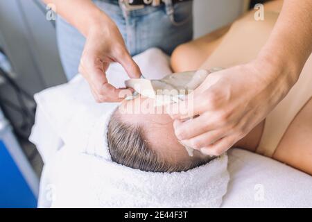Hoher Winkel der Ernte anonyme Kosmetikerin Entfernen Alginat Maske aus Gesicht der nicht erkennbaren weiblichen Klientin, die auf der medizinischen Couch liegt Schönheitssalon Stockfoto