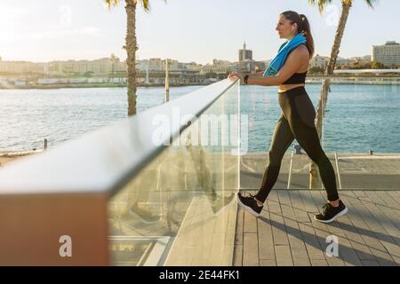 Ganzkörper-Seitenansicht der frauen Passform in schwarzer Sportbekleidung Strecken Sie Ihre Beine während des Fitnesstrainings am Wasser der Stadt Stockfoto