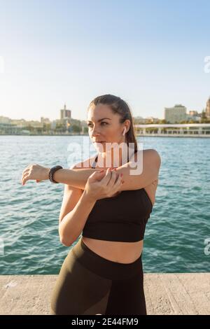 Selbstbewusste Erwachsene fit weiblich in schwarz Sportswear Stretching Arme während Übungen während des Outdoor-Trainings am Wasser Stockfoto