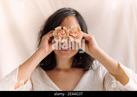 Lustige Porträt der lächelnden Frau mit rosa Gillyblumen auf den Augen und weißen Kleid und natürlichen Blick aus der Nähe. Freudig und glücklich Ausdruck mit Augen wi Stockfoto