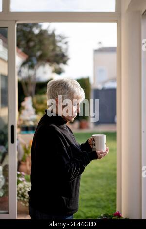 Ältere Frau in schwarz-weißem Haar, die im Winter zu Hause eine Tasse Kaffee am Fenster trinkt Stockfoto