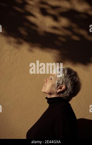 Seitenansicht einer entspannten älteren Frau an einer Wand mit geschlossenen Augen an einem sonnigen Tag Stockfoto