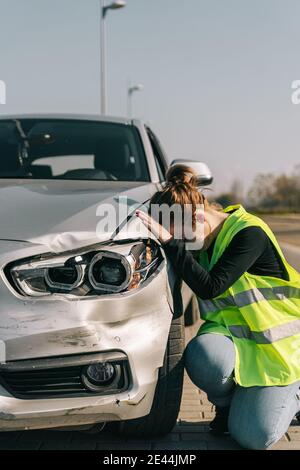 Seitenansicht der erlitt junge weibliche Fahrer in legerer Kleidung Und Verkehrssicherheitsweste lehnte Kopf auf beschädigte Autohaube Nach Unfall auf der Straße an sonnigen Tag Stockfoto