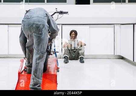 Schwarze weibliche Mechaniker mit Tablet und männliche Techniker Befestigung Motorrad Am Metallheber während der Arbeit in der Werkstatt Stockfoto