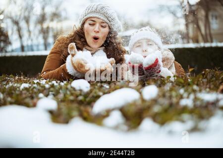 Happy stilvolle Mutter und Tochter in einem gestrickten Hüte und Schaffell Mäntel mit Fäustlingen in einem gestrickten Hut und Schaffell Mantel bläst Schnee draußen in der c Stockfoto