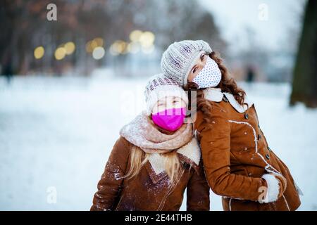Porträt von lächelnd stilvolle Mutter und Tochter in einem gestrickten Hüte und Schaffell Mäntel mit medizinischen Maske in einem gestrickten hut und Schaffell Mantel außen in Stockfoto