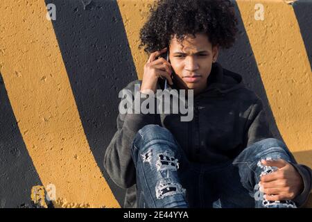 Serious afroamerikanischen männlichen Teenager in zerrissenen Jeans im Gespräch auf Smartphone, während Sie in der Nähe einer gestreiften Wand auf der Straße sitzen Stockfoto