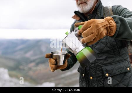 Cropped unkenntlich älteren männlichen Wanderer in Oberbekleidung in den Bergen stehen Im Winter und Gießen heißen Kaffee aus Geysir Kaffeemaschine in cup während VA Stockfoto