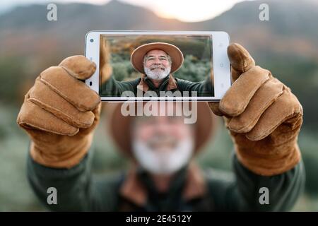 Lächelnder älterer männlicher Reisender in warmer Kleidung, der in den Bergen steht Und Selfie auf der Smartphone-Kamera während des Urlaubs in Caceres Stockfoto