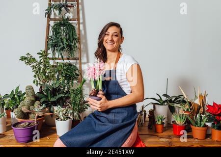Entzückende Gärtnerin in Schürze sitzt auf Holztisch mit Verschiedene Topfpflanzen im Blumenladen und Blick auf die Kamera Stockfoto