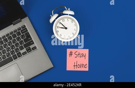 Stay Home Flat Lay Workplace Konzept mit einer Textbeschriftung, Laptop und Wecker auf einem blauen Hintergrund Foto Stockfoto