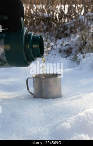Im Winter gießt ein Mann heißen Tee aus einer Thermoskanne in einen Becher am Fluss. Konzept der Winter Outdoor-Erholung. Speicherplatz kopieren. Vertikale Ausrichtung. Stockfoto