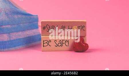 Stay Home Konzept mit einer medizinischen Maske auf einem rosa Background, mit Holzblöcken, Tetxt Inschrift und rotem Herz, valentinstag Urlaub Foto Stockfoto