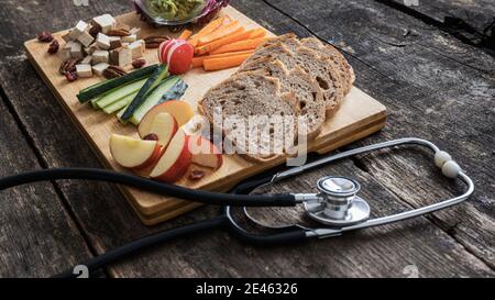 Gesunde Ernährung konzeptuelles Bild - hölzerner Servierteller mit Gemüse, Fruchtsauerteigbrot und veganem Protein neben einem Stethoskop. Stockfoto