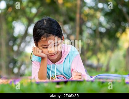 Das asiatische Mädchen legte sich hin und legte ihre rechte Hand unter das Kinn auf das Buch mit einem Lächeln auf den Rasen im Garten. Das Kind liest ein Buch auf dem Stockfoto