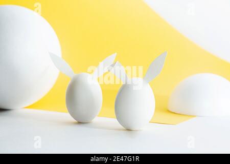 Minimalistisches Osterkonzept auf gelb-weißem Hintergrund. Weiße Ostereier mit Hasenohren und weißen geometrischen Formen. Stockfoto