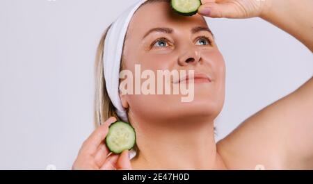 Nahaufnahme einer älteren Frau mit gepflegter, strahlender Haut und einem süßen Lächeln. Hält Gurkenscheiben und blickt auf einen weißen Hintergrund. Stockfoto