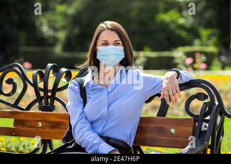 Schöne junge Frau sitzt auf einer Sommerparkbank und trägt eine schützende medizinische Gesichtsmaske. Stockfoto