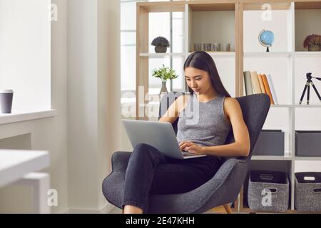 Junge positive asiatische Frau Büroangestellte sitzen in Sessel mit Laptop und Online-Arbeit Stockfoto