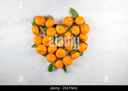 Orangefarbene Mandarinen mit grünen Blättern in Herzform auf weißem Steingrund. Herz aus Mandarinen. Blick von oben. Speicherplatz kopieren. Oben Stockfoto