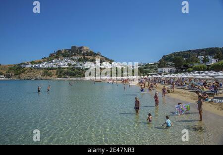 RHODOS, GRIECHENLAND - Apr 02, 2020: Ein sonniger Tag am Lindos Strand an der warmen Ägäis. Im Hintergrund das Dorf Lindos und darüber Lindos cas Stockfoto