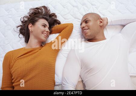 Draufsicht Aufnahme eines glücklichen multiethnischen Paares, das zusammen auf orthopädischem Bett liegt. Junges Paar lächelt sich an, liegt auf einem neuen Bett bei Möbel stor Stockfoto