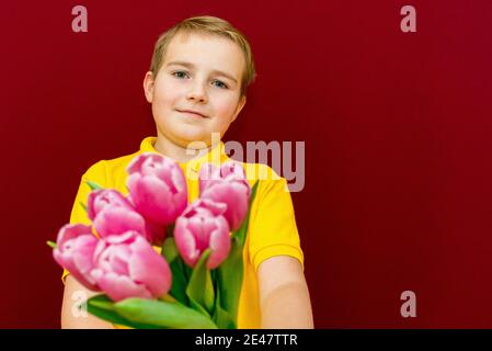 Junge geben Blumen Bouquet, Kind hält lila Tulpen Bündel, Blick auf Camera.Studio rot oder burgunderrot Hintergrund. Stockfoto