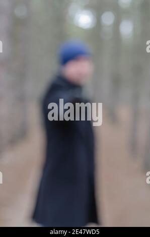 Unscharfe, unscharfe Darstellung eines Mannes, der mit einem Gewehr auf die Kamera zeigt. Mann in einem schwarzen Mantel und blauen Hut zeigt seine rechte Hand mit einem Gewehr direkt auf Th Stockfoto
