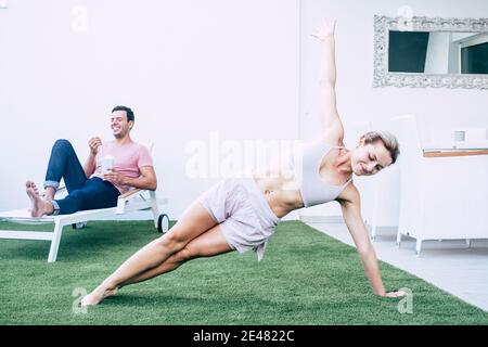 Fitness Sport Aktivität zu Hause mit schönen Mädchen tun Pilates Übungen im Garten, während sich der Boyfroend auf der Sonnenliege entspannt - junge Frau in gesund Stockfoto