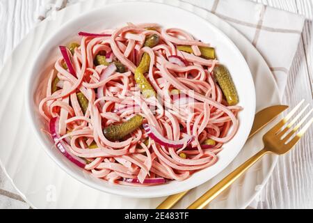 Deutscher Wurstsalat aus geschnittener lyoner-Wurst mit Gurken und Rote Zwiebel serviert auf einem weißen Teller mit goldenem Besteck Und das Roggenbrot auf dem weißen Holz Stockfoto