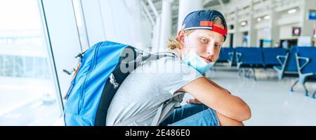 Porträt eines einsamen Teenager-Alleinreisenden mit Rucksack in leerer Flughafen-Passagiertransferhalle in schützender Gesichtsmaske und trauriger Betrachtung der Kamera. Tr Stockfoto