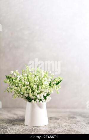 Blumenstrauß von Lilien des Tales in einem weißen Glas auf einem grauen Betongrund. Stockfoto