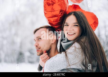 Junges Paar in der Liebe im Park auf einem Hintergrund von Schnee und roten herzförmigen Ballons. Geschenk zum Valentinstag. Die Frau sitzt auf dem Rücken des Mannes. Stockfoto