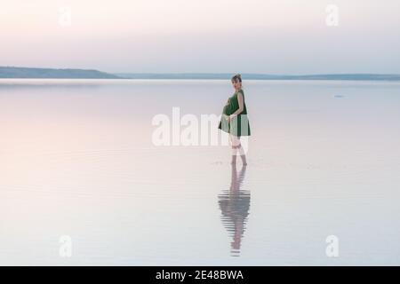 Schöne schwangere Frau, die im Wasser in den Strahlen der untergehenden Sonne steht. Wunderbarer Abend am Strand Stockfoto