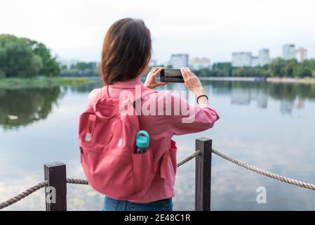 Nahaufnahme, ein Blick von hinten, ein Mädchen im Sommerteich des Flusssees, zeichnet Videos und Fotos auf einem Smartphone auf. Besichtigungstouren. Landschaftsfotografie Stockfoto