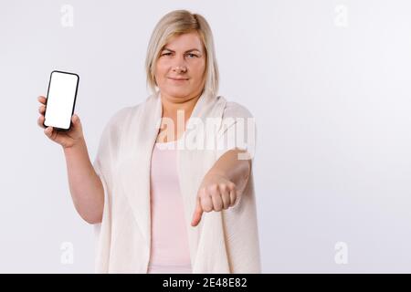 Unzufriedene kaukasische Frau zeigt einen Telefonbildschirm und mit der anderen Hand ein Abneigung Zeichen. Daumen nach unten auf weißem Hintergrund. Stockfoto