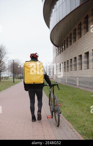 Vertikale Rückansicht eines Kurierdienstes mit Thermo-Lieferung Rucksack zu Fuß in der Stadt mit seinem Fahrrad Stockfoto
