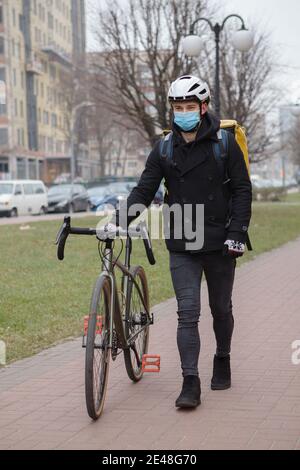 Männlicher Kurier trägt medizinische Gesichtsmaske, geht mit seinem Fahrrad, trägt Thermorucksack Stockfoto