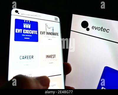 Person, die Smartphone mit Website des deutschen Biotechnologie-Unternehmens Evotec SE neben Computer-Monitor hält. Konzentrieren Sie sich auf das Unternehmenslogo auf dem rechten Bildschirm. Stockfoto