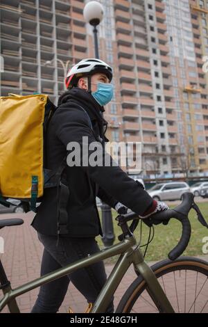 Vertikale Aufnahme eines Kuriers in medizinischer Gesichtsmaske, der mit seinem Fahrrad unterwegs ist und sich in der Stadt umsieht Stockfoto
